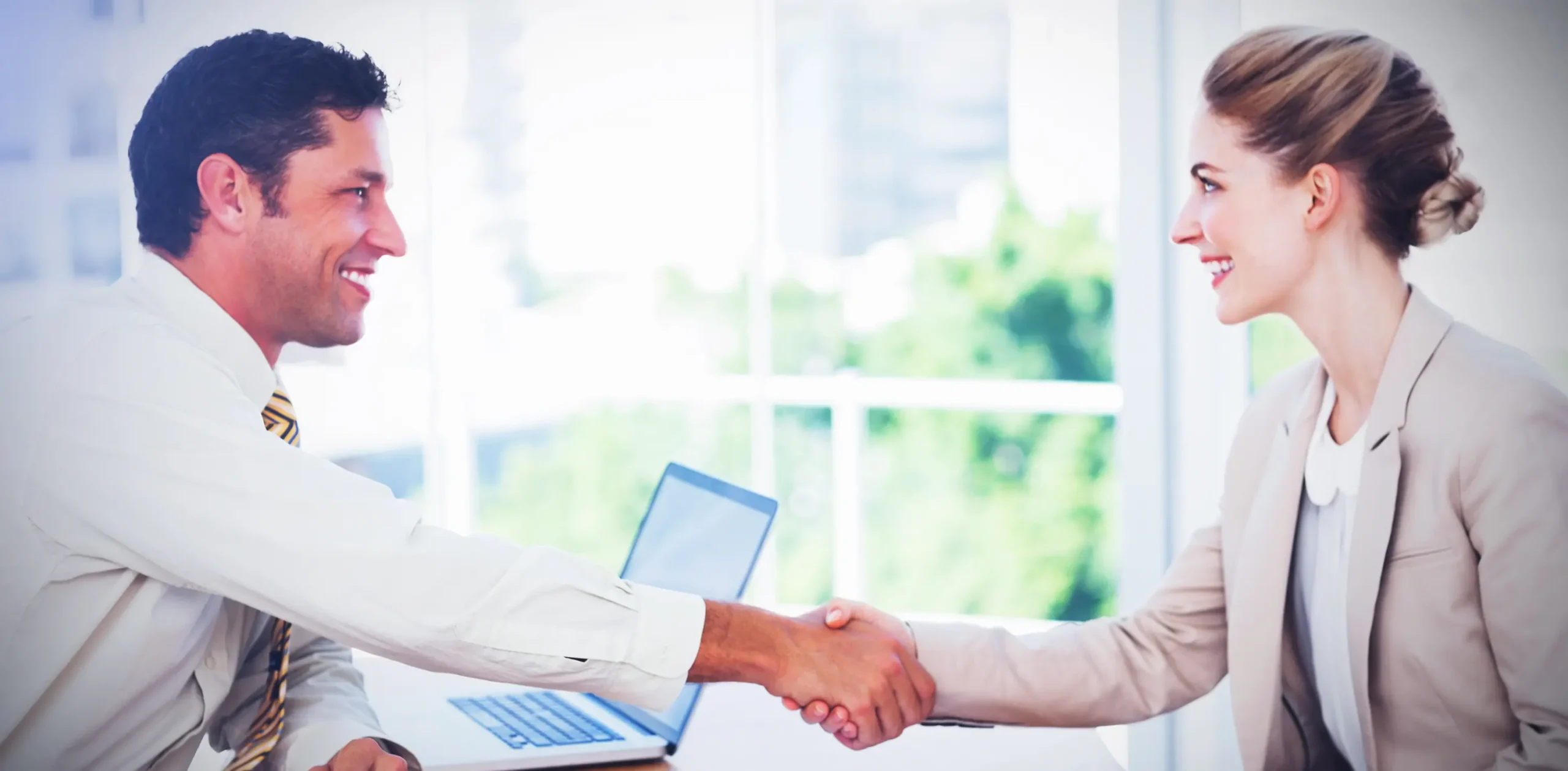 A smiling man and woman in business attire warmly shaking hands across a desk with a laptop, in a bright office setting. Symbolizes the welcoming collaboration with our new branding partner — professionals who believe great partnerships begin with strong connections and clear, confident communication.