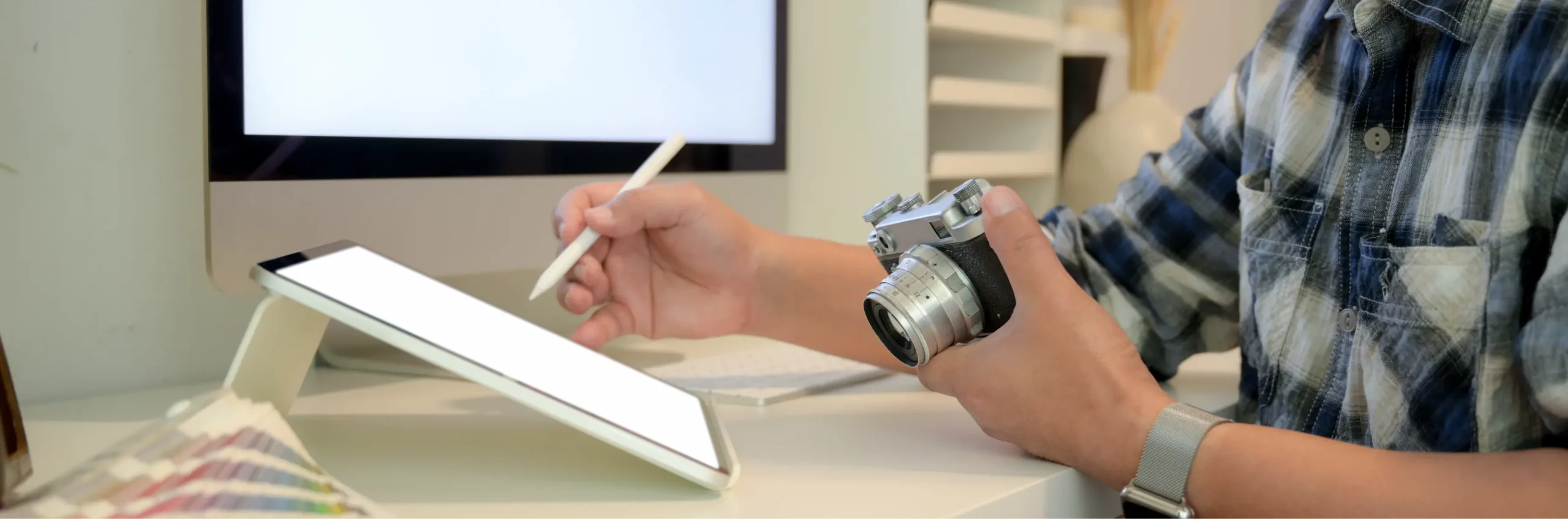 A creative professional holding a vintage-style camera in one hand while sketching on a digital tablet with a stylus, with a computer screen and color palette in the background. Represents our new branding partner’s hands-on, design-led approach — blending sharp visuals, smart content, and authentic storytelling in every brand they shape.