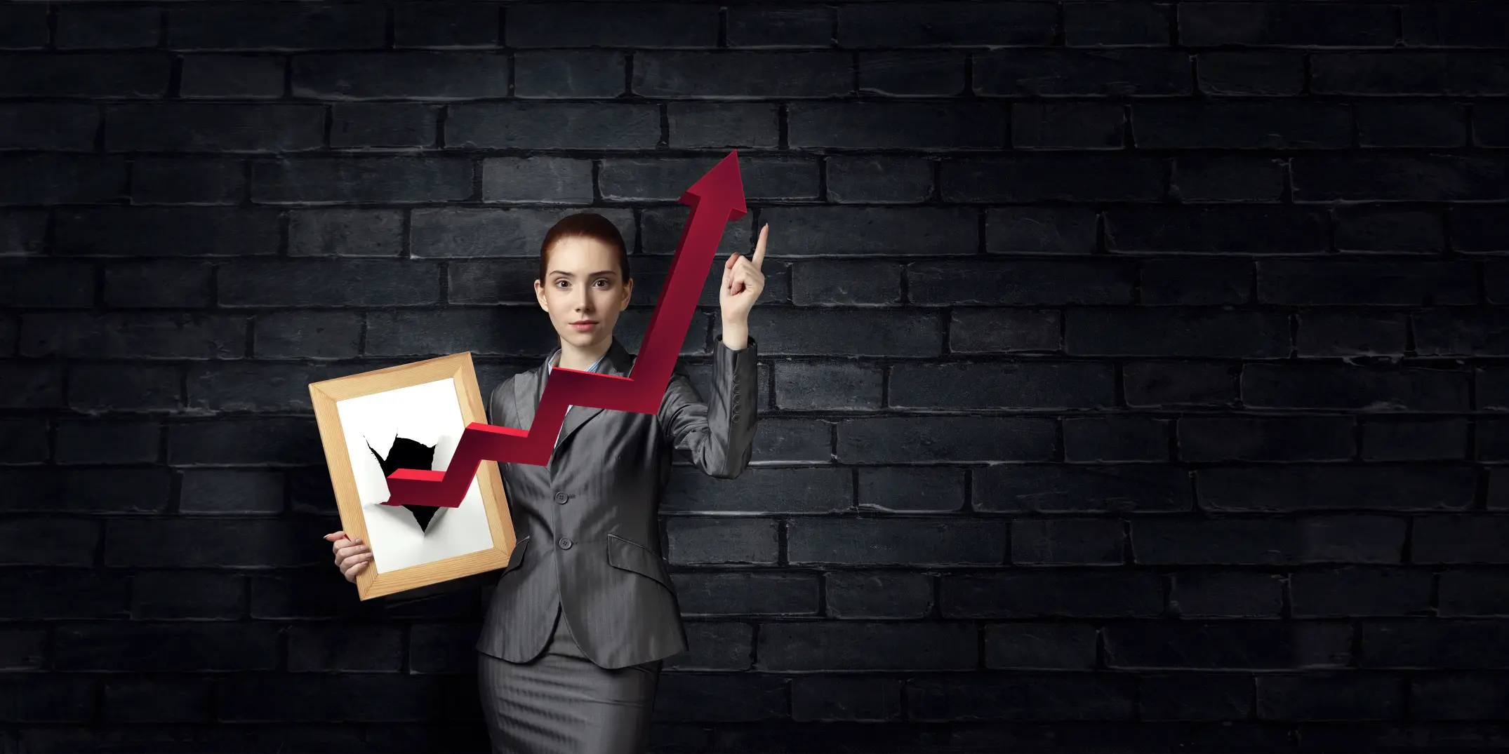 A poised businesswoman in a grey suit stands against a dark brick wall, holding a picture frame with a red arrow breaking through and rising upward. Symbolizes our new branding partner’s bold, no-limits mindset in driving brand visibility and growth.